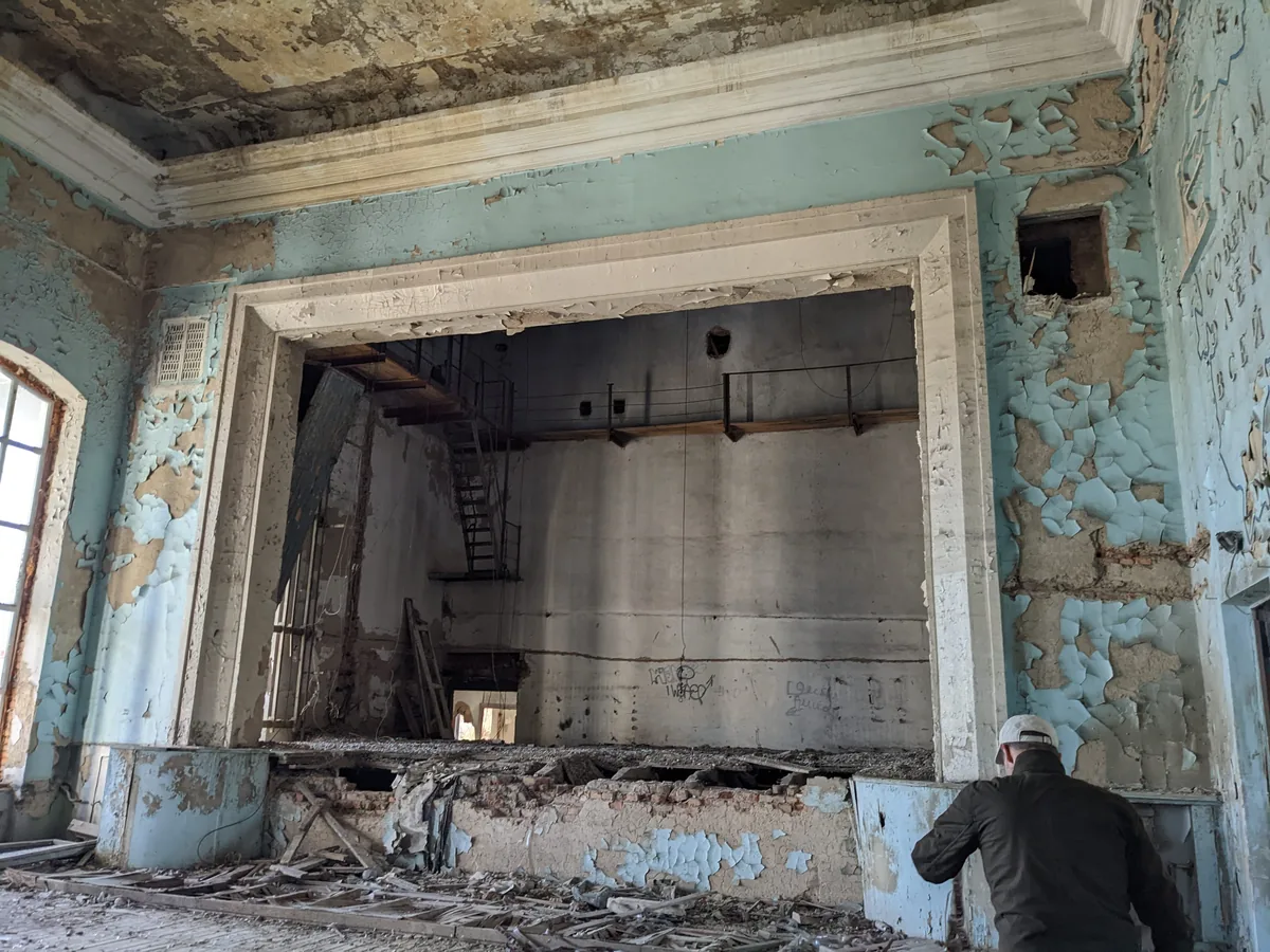 Interior of abandoned Soviet theatre with peeling paint and collapsed stage, visitor with hard hat
