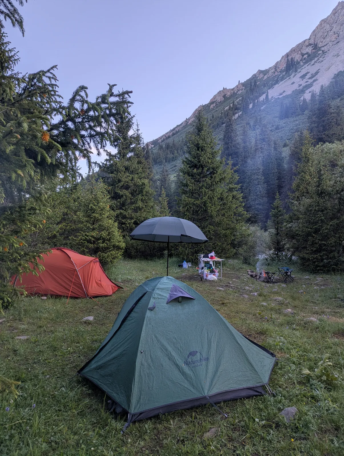 A camp at dusk with two tents, a campfire still going, a steep mountain rising behind